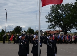 Ochotnicy z Mistowa podnoszą flagę państwowa. W tle widać strażaków i poczty sztandarowe