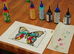 Kolorowy motyl namalowany na szkle. Obok rysunku stoją farby