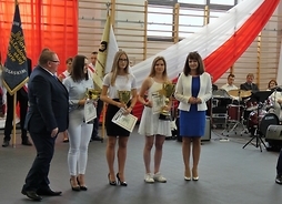 Najcelniejsze uczennice w kategorii pistolet pneumatyczny stoją trzymając puchary, obok wicemarszałka Janiny Ewy Orzełowskiej. W tle widać poczty sztandarowe i flagę państwową.