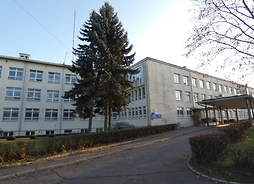 Zdjęcie przedstawia budynek szpitala. Po prawej stronie widać podjazd dla karetek. Przy budynku rosną świerki