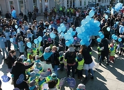 Na ulicy, w dużym planie - dziesiątki uczestników niebieskiego korowodu, dzieci i młodzież z placówek szkolnych i przedszkolnych z wychowawcami - niosą niebieskie balony, kartonowe niebieskie serca.