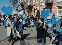 Ulicą maszerują radosnie uczestnicy korowodu - w dużym planie widać dziesiatki uczestników, dzieci i młodzież ze szkół i przedszkoli z wychowawcami - niosą niebieskie balony, kartonowe niebieskie serca i emblematy swoich placówek.