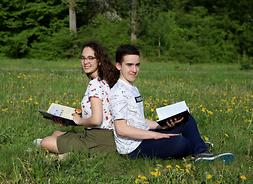 Dwoje młodych uczniów, dziewczyna i chłopak, siedzą z otwartymi książkami w rękach, plecami do siebie, na trawie.