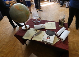 Na biurku leżą stare księgi, zeszyty, dokumenty. Z boku biurka stoi zabytkowy globus