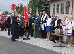 ilkoro ludzi stoi w rzędzie na nwym chodniku. Wśród nich jest członek zarządu Janina Ewa Orzełowska. Po prawej stronie stoi stolik z nożycami, po lewej flagi