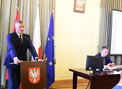 Przy mównicy z godłem województwa przemawia wicemarszałek Rafał Rajkowski. Ręce ma oparte o pulpit. Na prawo widać stolik, przy którym siedzi przewodniczący sejmiku