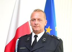 druh w mundurze strażackim pozuje wyprostowany do zdjęcia, ręce ma opuszczone wzdłuż tułowia, za nim stoją flagi Polski i UE