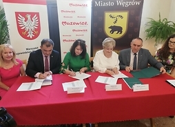 Przy stole prezydialnym zakrytym obrusem siedzi sześć osób  beneficjentów z Węgrowa. Elżebieta Lanc i Janina Orzełowska siedzą w środku i podpisują dokumenty. W tle banery z herbem Mazowsza, herbem Węgrowa i logotypami