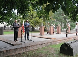 Dwaj mężczyźni w garniturach stoją przy mikrofonach, w tle pomniki osób straconych oraz uczestnicy uroczystości.
