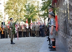 Przed wysokim marmurowym pomnikiem stoją na baczność harcerze i żołnierze, na przeciw nich stoi żołnierz z wieńcem biało-czerwonym.