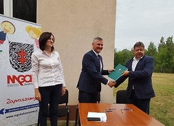 Przy kwadratowym stoliku ustawonym na zewnątrz nudynku stoi wicemarszałek i podaje zieloną teczkę mężczyźnie w garniturze. Obaj się uśmiechają i patrzą z głową zwróconą do kamery. Obok nich stoi kobieta, tak samo się uśmiecha