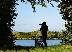 mężczyzna stoi nad jeziorem, robi zdjęcie, oboik siedzi pies, wokół widać las