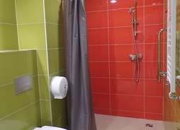 Łazienka szpitalna. Na wprost - nowy prysznic z zasłoną, obok niego WC. Ściany od podłogi do sufitu są wyłożone jednokolorowymi płytkami. Wszystko lśni nowowścią