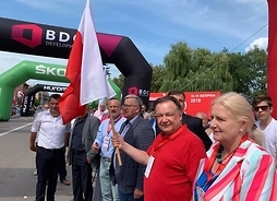 marszałek z flagą Polski stoi przy krawędzi jezdni uśmiecha się