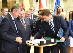 Przy stoliku stoi prezydent Warszawy. Pochyla się nad zieloną teczką i podpisuje deklarację. Otaczają go inni sygnatariusze