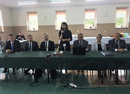 Widok na stół konferencyjny, przy którym siedzi siedem osób, po środku Janina Ewa Orzełowska, która stoi i trzyma w ręku mikrofon.