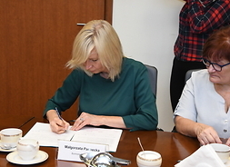 Kobieta o blond włosach siedzi przy stole. Pochyla głowę nad dokumentem, który właśnie podpisuje