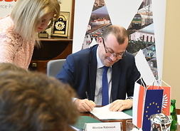 Przy stole siedzi wicemarszałek i podpisuje dokument. Przed nim są proporczyki z flagą Polski i UE