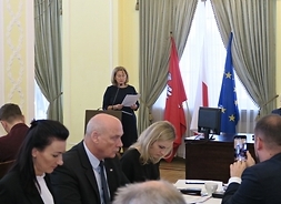 Na zdjęciu widać część stołu, przy którym siedzą radni, na drugim planie jest mównica, przy której stoi koieta. Obok mównicy widać trzy flagi - z herbem Mazowsza, flagę Polski i UE