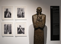 Po prawej stronie widać rzeźbę stojącego człowieka z pochyloną głową i złożonym jak do modlitwy rękami. Obok na ścianie widać cztery czarnobiałe zdjęcia. Dwa z nich przedstawiają człowieka z podobną pozą.