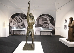 Zdjęcie sali z wystawą. Na środku stoi rzeźba człowieka z uniesioną do góry ręką. W tle widać dwie półokrągłe nawy z rzeźbami. Po prawej stronie przy ścianie też widać rzeźbę