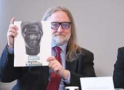 Dyrektor Klejnocki (pan z brodą, okularach i długimi do ramion włosami) trzyma obok swojej twarzy książkę