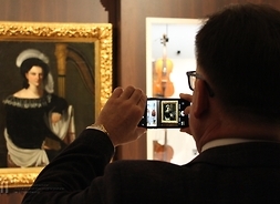 Marszałek stoi tyłem i unosi ręce, robiąc zdjęcie portretowi damy. W monitorze telefonu widać obraz, któremu marszałek robi zdjęcie