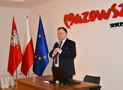 Marszałek stoi za stołem, trzymając w obu dłoniach mikrofon. Po jego prawej stronie widać flagi UE, Polski i województwa. Z tyłu na ścianie jest napis Mazowsze serce Polki