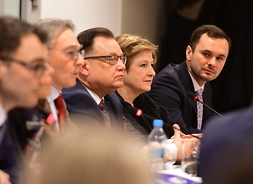 Widok z boku na siedzących przy głównym stole przedstawicieli samorządu i komisji europejskiej. Ostatni mężczyzna przy stole jest pochylony do przodu i patrzy prosto na kamerę