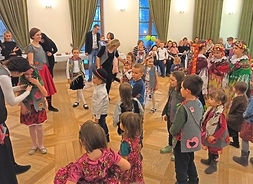 Kilkunaścioro dzieci i ich rodzice oraz opiekunowie wykonują figury taneczne na dużej sali.