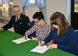 Na pierwszym planie przy stole siedzi Janina Ewa Orzełowska, za nią kobieta w okularach oraz mężczyzna w garniturze z długopisem w prawym ręku, którą opiera o blat. Obie panie podpisują dokumenty