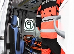 Wnetrze ambulansu, widać elementy wyposażenia: nosze transportowe, transporter - tyłem widoczny ratownik w kombinezonie.
