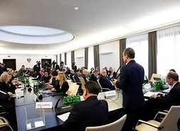 Widek zza stołu prezydialnego na salę obrad, stoi tyłem do obiektywu marszałek Tomasz Grodzki, za stołami siedza uczestnicy konferencji.