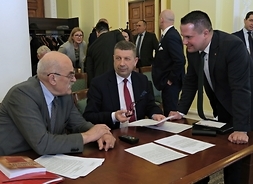 Dwóch mężczyzn siedzi przy stole, na którym są rozłożone dokumenty. Trzeci mężczyzna stoi przy stole i pochyla się lekko w ich kierunku
