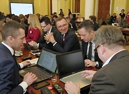 Zdjęcie pokazuje róg stołu. Dwóch radnych ma otworzone laptopy, ręce trzymają ma klawiaturze. W tle widać inne stoły z radnymi