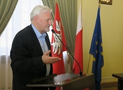 Starszy pan stoi bokiem przy mównicy. W tle widać flagi Polski, UE i województwa