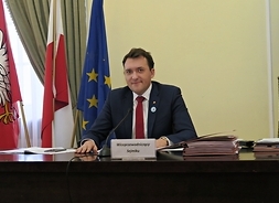 Za dużym biurkiem siedzi mężczyzna w garniturze i krawacie. Patrzy przed siebie, po jego prawej stronie stoją trzy flagi: Polski, województwa i UE