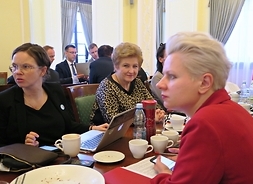 Na pierwszym planie widać dwie kobiety siedzące naprzeciw siebie i patrzące w lewo. Obok jednej z nich siedzi Elżbieta Lanc, która patrzy w kamerę