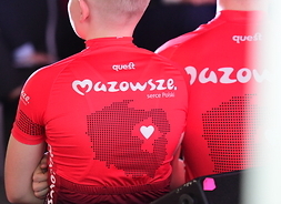 Dwóch zawodników widocznych od tyłu w pełnym planie, od góry do pasa, ubranych w koszulki czerwone z logotypem Mazowsza.