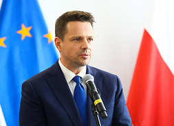 Prezydent m.st. Warszawy Rafał Trzaskowski stoi przed mikrofonem, za nim flagi Polski i Unii Europejskiej.