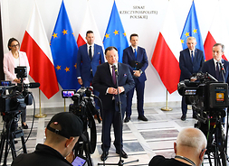 W dużym planie siedmioro uczestników konferencji stojących w 2 rzedach z odstepami. Za nimi flagi Polski i Unii Europejskiej.