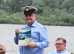 Marszałek w marynarskiej czapce z daszkiem stoi na tle rzeki i pokazuje książkę pt.