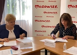 Elżbieta Lanc i Janina Ewa Orzełowska siedząc podpisują umowy.