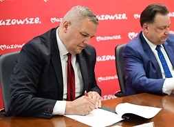 Dyrektor Lewandowski siedzi za stołem. Pochyla się nad dokumentem, który podpisuje. Po jego lewej stronie siedzi marszałek.