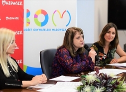 Trzy kobiety siedzą za stołem, siedząca w środku mówi do mikrofonu.