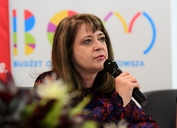 Janina Ewa Orzełowska siedzi za stołem i mówi do mikrofonu.