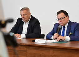 Marszałek Struzik i dyrektor Lewandowski siedzą za stołem konferencyjnym obok siebie. Marszałek trzyma w ręku mikrofon. Przed nim jest plakietka z jego nazwiskiem