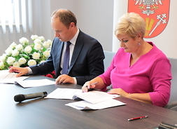 Starosta Zalewski i wicestarosta Beata Jóźwiak siedzą przy stole i podpisują dokumenty. W tle widać baner z herbem Mazowsza