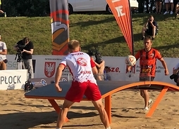 Dwóch zawodników - -jeden z nich to wicemistrz Polski Duszak - gra przy stole na plaży. Nad sołem widać piłkę nożną w locie.