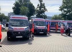 Pięć ambulansów stojących na placu w układzie wahlarzowym, przed nimi ratownik, po prawej Elżbieta Lanc przed mikrofonem obk której dyrektor firmy.